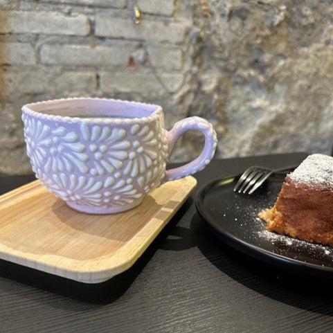 Ceramic mug with floral pattern on a wooden coaster next to a plate of cake on a stone surface.